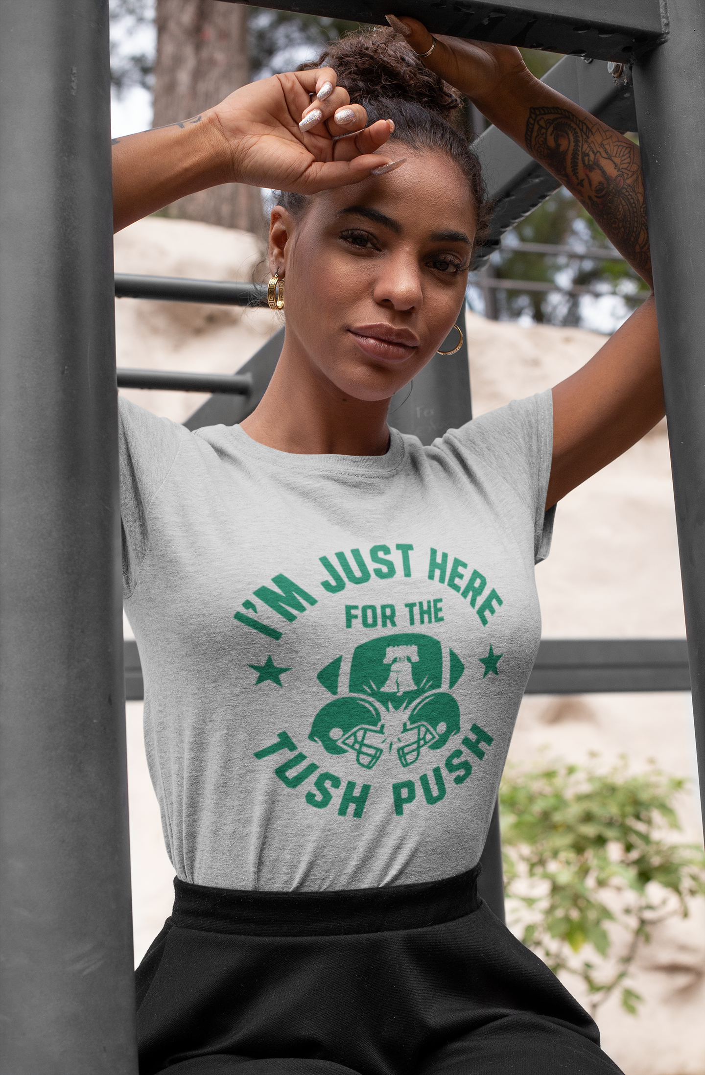 Woman wearing a gray tush push t-shirt with green text and graphic, sitting outdoors.