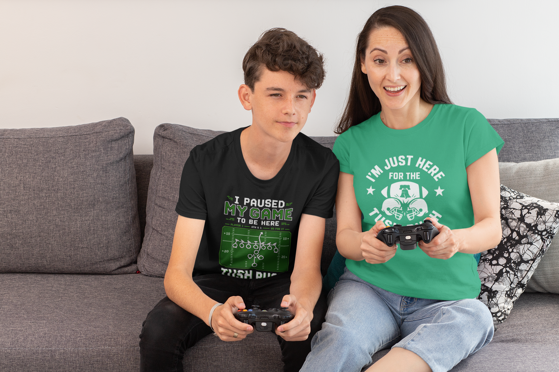 Two people sitting on a couch, one wearing a black t-shirt with gaming text holding a controller and the other in a green t-shirt holding a controller.