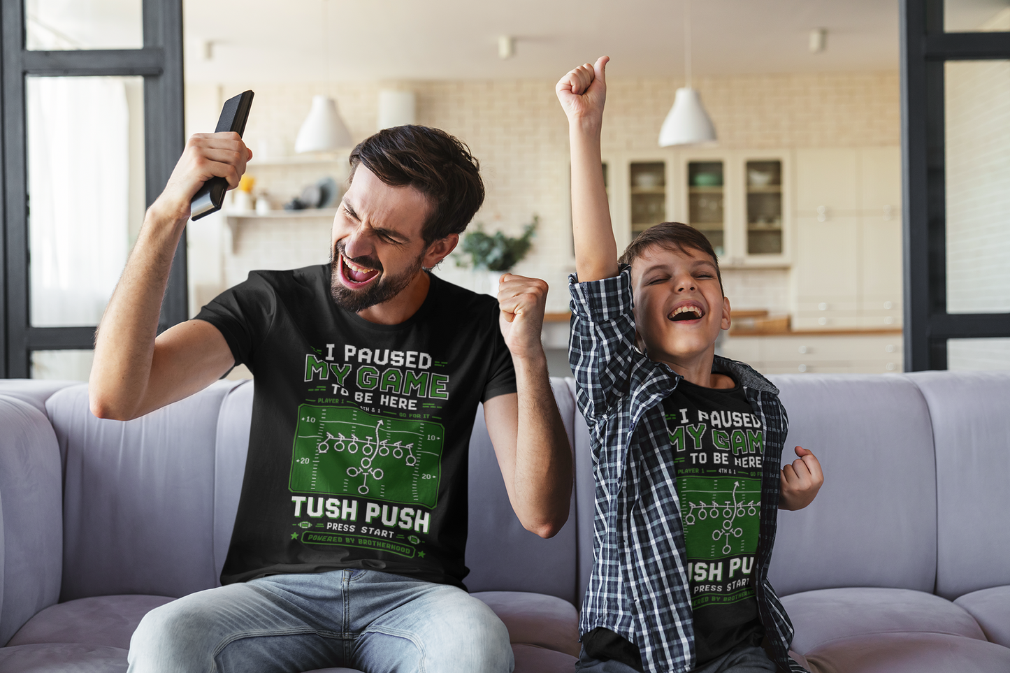 Man and child sitting on a couch with matching t-shirts, celebrating in a living room.